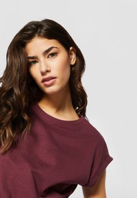 Bordeaux t-shirt med kort ærme og rund halsudskæring, blød tekstur og opkrøllede ærmer, båret af en person med langt, bølget hår.