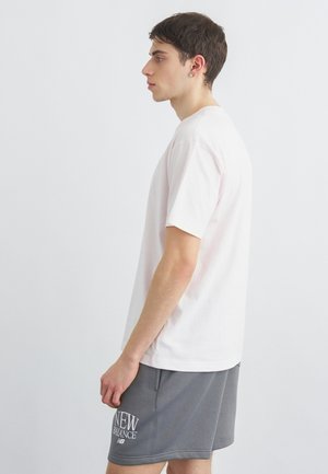 Jaunas vyras dėvi baltą trumpomis rankovėmis marškinį ir pilkas New Balance šortų, stovintis šonu prieš paprastą baltą foną.