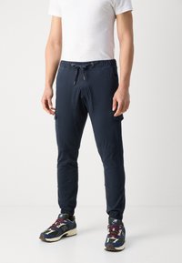 Man in een donkerblauwe jogger broek, witte t-shirt en multicolor New Balance sneakers, staande tegen een egale witte achtergrond.