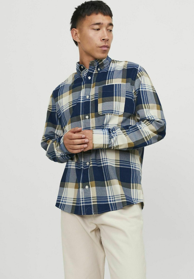 Camisa de cuadros que presenta una mezcla de colores azul marino, beige y verde oliva. Tiene un diseño de botones, un cuello y un bolsillo en el pecho.