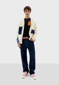 Cárdigan de punto crema con parches azules y oscuros, camiseta negra con un gráfico colorido, vaqueros oscuros y zapatillas rosas con suelas blancas.