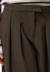 Rjave hlače s pliseji in gladko teksturo, z ravnim sprednjim delom, stranskimi žepi ter detajlom na pasu. Nad njimi je viden črtast srajčni zgornji del.