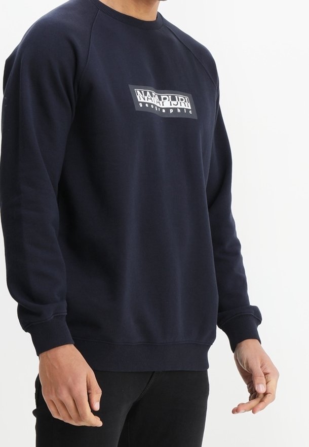 Homme portant un sweat-shirt bleu marine avec un logo rectangulaire blanc mentionnant "Napapijri Geographic" sur la poitrine, avec un pantalon noir.