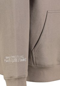 Pullover-Hoodie in hellem Taupe, aus weichem Stoff. Verfügt über eine Kängurutasche und weißen gestickten Text auf dem Ärmel.