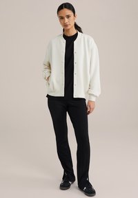 Jachetă bomber albă cu nasturi tip snap, purtată peste un tricou negru, asortată cu pantaloni negri și adidași negri. Țesătură netedă, neîmbelșugată.
