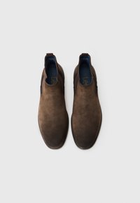 Botas marrones de ante sin cordones con paneles laterales elásticos y tiradores de tela, mostradas desde una vista superior sobre un fondo blanco liso.