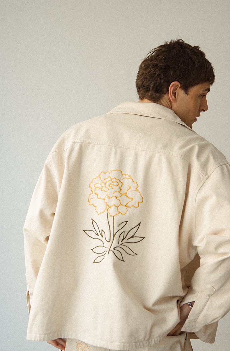 Veste beige avec une grande broderie florale jaune au dos. Le tissu semble doux, avec une coupe ample et décontractée ainsi que des détails de couture visibles.