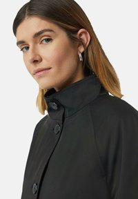 Schwarze Jacke mit hohem Kragen, ausgestattet mit zwei runden Knöpfen und einer glatten, matten Textur. Das Haar ist glatt und schulterlang.