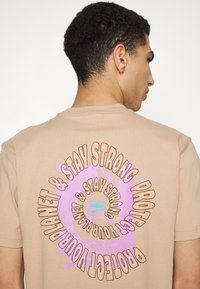 T-shirt en coton beige à manches courtes, avec un design graphique en violet et noir au dos, texte disposé en motif circulaire.