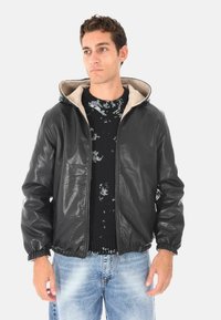 Uomo con giacca di pelle nera con cappuccio sopra un maglione nero usurato e jeans chiari blu sbiaditi, in piedi contro uno sfondo bianco.