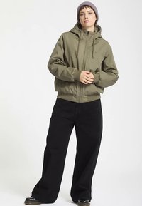 Giacca verde oliva con cappuccio, chiusura con zip e polsini a coste, abbinata a pantaloni neri a gamba larga. Il modello indossa un berretto viola chiaro.