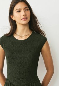 Ciemnozielona sukienka o fakturowanym materiale, z krótkimi rękawami i dopasowanym gorsetem. Wykończenie stanowi złoty łańcuszek z małymi ozdobami.