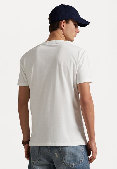 Tricou alb din bumbac cu guler rotund și mâneci scurte, purtat cu blugi din denim ușor deteriorați, de culoare albastru deschis, și o șapcă de baseball bleumarin.