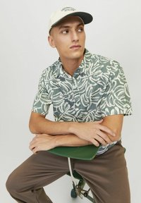 Camicia a maniche corte con un motivo a foglie verde e bianco, tessuto con una texture liscia. Pantaloni marroni e un cappellino beige con scritte verdi.