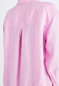 Camicia di lino rosa chiaro con una piega sul retro e colletto, con maniche ampie e una texture morbida. Il materiale liscio cade delicatamente.