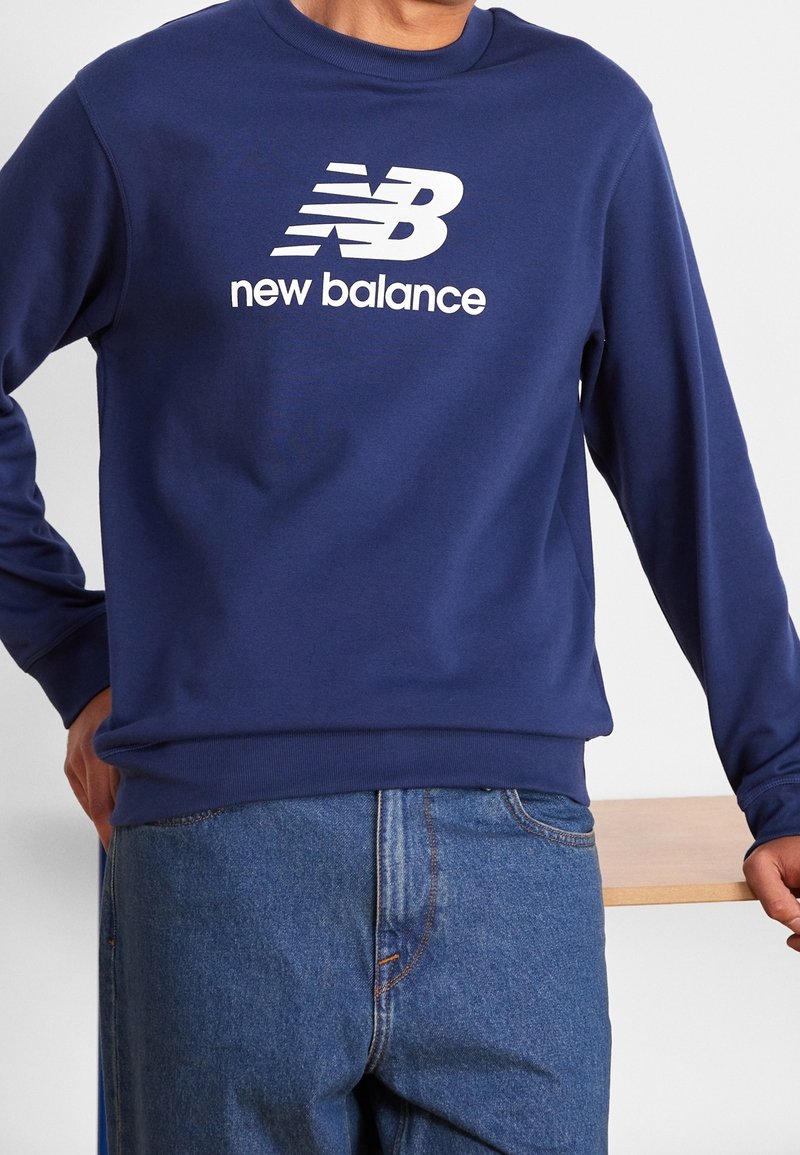 Personne portant un sweat-shirt New Balance bleu marine avec un logo et un texte blancs, associé à un jean en denim bleu, s'appuyant sur une surface en bois.