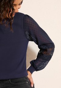 Tumesinine kampsun soonikkoes kehaga ja läbipaistvate polka dot varrukatega, mis on kaunistatud pitsi detailide ning elastsete mansettidega. Lihtne, keha järgiva lõikega disain õrnade detailidega.