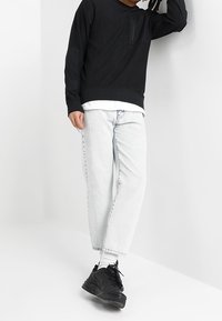 Sudadera negra con un bolsillo frontal, combinada con jeans holgados de color azul claro y zapatillas negras, llevadas con calcetines blancos.