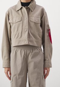 Osoba w beżowej, krótkiej kurtce oraz dopasowanych, spodni wysokiego stanu, z czerwoną metką przymocowaną do zamka w lewym kieszeni rękawa.