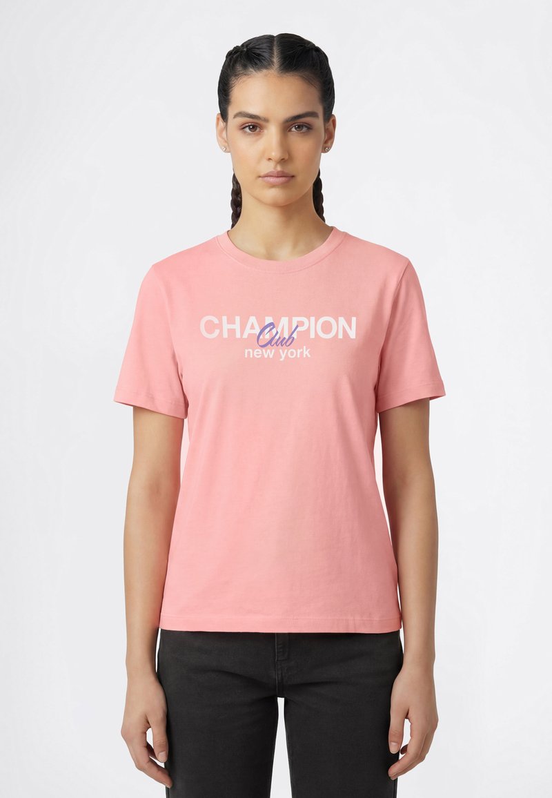 Mujer con cabello trenzado que lleva una camiseta rosa con el texto "Champion Club New York" y pantalones negros, posando frente a un fondo liso.
