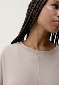 Felpa beige chiaro con scollo rotondo, texture liscia e dettagli di cucitura sottili sulla parte anteriore. I capelli intrecciati sono visibili accanto.