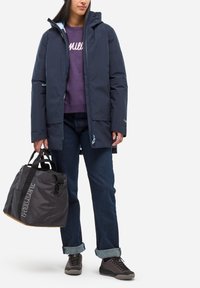 Granatowa wodoodporna parka z kapturem, noszona na fioletowej bluzie. Ciemne dżinsy i szare buty do wędrówek. Czarna torba sportowa z logo.