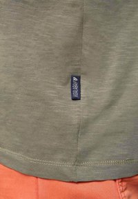 Primer plano de tela verde oliva con una pequeña etiqueta negra cosida cerca del dobladillo, combinada con pantalones naranjas debajo.