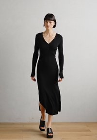 Black ribbed knit dress with long sleeves, deep v-neck, button detail, and side slit, paired with black platform sandals.