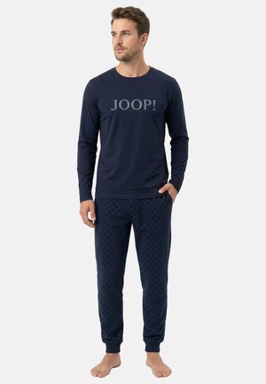 Man staat op blote voeten, draagt een marineblauw shirt met lange mouwen waarop "JOOP!" staat, en een marineblauwe joggerbroek met patroon tegen een effen achtergrond.
