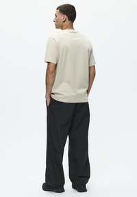 Beige bomulds t-shirt med korte ærmer, rund halsudskæring og afslappet pasform, parret med løse sorte bukser og sorte fodtøj. Minimalistisk design.