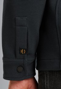 Temno obarvana teksturirana jakna z vafljastim vzorcem, ki vsebuje blagovno oznako in detajl gumbov na manšeti, kar poudarja vzdržljivost in oblikovanje.