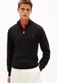 Μαύρο πλεκτό πουλόβερ με φερμουάρ μέχρι τον λαιμό και ριμπ υφή καθώς και γιακά. Διαθέτει μικρό λογότυπο στο στήθος. Φορεμένο με ανοιχτόχρωμο παντελόνι.