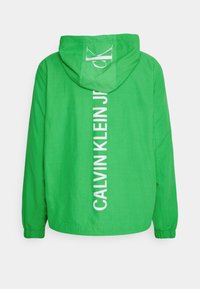 Groene hoodie gemaakt van lichtgewicht stof. Heeft witte tekst "CALVIN KLEIN JR" verticaal langs de achterkant. Casual pasvorm met elastische manchetten.