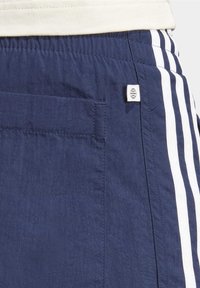 Námořnické modré šortky s bílým páskem v pase, které mají bílé pruhy po stranách, zadní kapsu a malou značka. Materiál vypadá lehce a texturovaně.