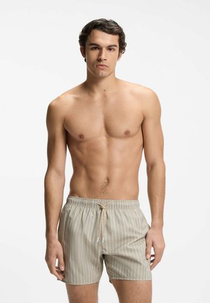 Young man standing, shirtless, wearing beige striped swim shorts with a drawstring, against a plain white background.