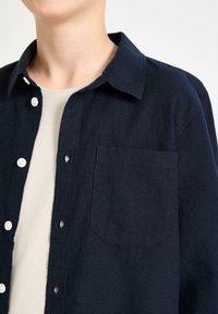 Camicia blu navy con bottoni in un tessuto a trama, dotata di colletto a punta, una tasca sul petto e leggeri accenti sui bottoni. Strato interno morbido visibile.