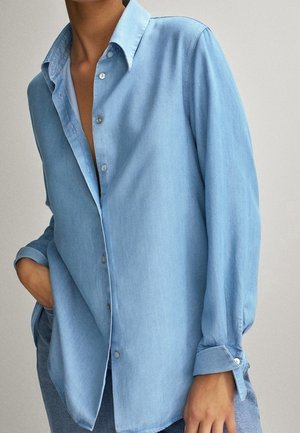 Personne portant une chemise en denim bleu clair à boutons, manches retroussées, une main dans la poche, debout devant un fond uni.