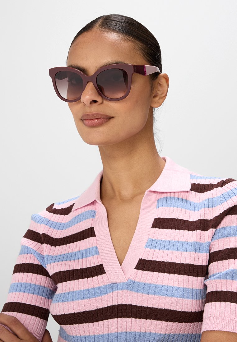 Lunettes de soleil violettes en forme d'yeux de chat, avec des verres foncés et une monture brillante. Les branches présentent un accent contrastant subtil.