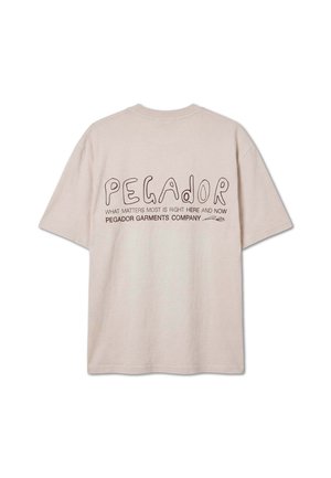 Maglietta beige a maniche corte con "PEGADOR" in grandi lettere e il testo "What matters most is right here and now" sul retro.
