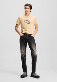 Camiseta de algodón beige con estampado negro, combinada con jeans de mezclilla gris oscuro con efecto deslavado. Botines negros completan el look.