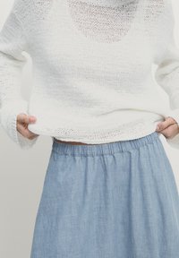 Persona che indossa un maglione di lana bianco e una gonna azzurro chiaro, mentre sistema il maglione in vita su uno sfondo semplice.