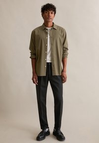 Koszula khaki zapinana na guziki, noszona na białej koszulce, w połączeniu z czarnymi spodniami i czarnymi mokasynami, o luźnym kroju i podwiniętych rękawach.