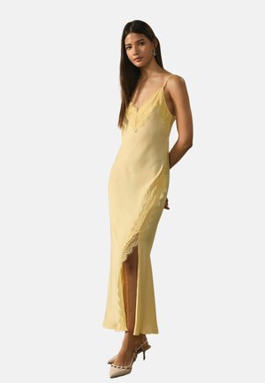 Woman wearing a sleeveless yellow slip dress with lace trim and side slit, paired with beige pointed heels, standing with hands behind back.