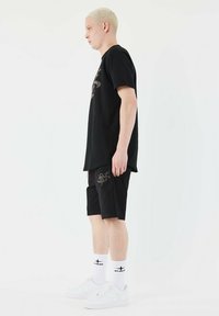 Black cotton T-shirt with a large silver logo, paired with matching black shorts featuring a small design. White sneakers and socks complete the look.