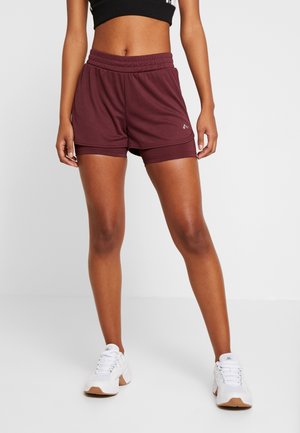 Femme portant un short de sport bordeaux à double couche, un soutien-gorge de sport noir et des baskets blanches, debout devant un fond uni.