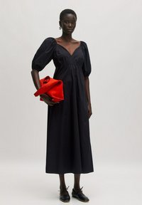Schwarzes Maxikleid mit Puffärmeln und V-Ausschnitt, aus glattem Stoff. Hält eine rote gefaltete Clutch-Tasche und kombiniert mit schwarzen flachen Schuhen.