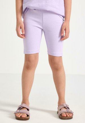 Dziecko noszące jasnofioletowe krótkie spodenki do kolan oraz pasujące sandały, stojące na jednolitym białym tle.