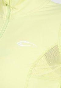 Haut de sport jaune clair avec un logo panthère stylisé blanc et des panneaux de ventilation en mesh près de l'épaule.