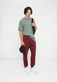 Mlad moški, oblečen v zeleno majico Nike, maroon trenirke z belimi črtami, bele teniske, drži črn klobuk in torbo preko rame na belem ozadju.