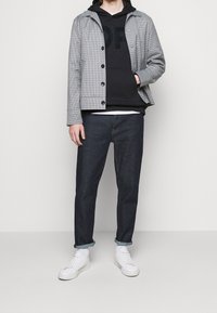 Chaqueta a cuadros gris sobre una sudadera negra con capucha, jeans de mezclilla oscuros y zapatillas blancas. La chaqueta cuenta con un cierre de botones y una silueta casual.